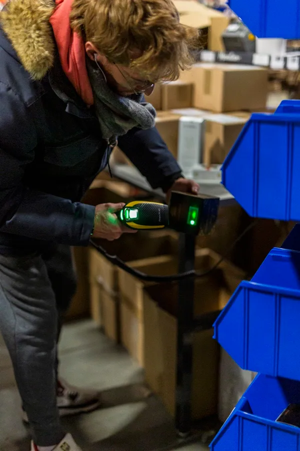 Opérateur scannant un article au lecteur de codes-barres, bacs de picking bleus, cartons en attente.