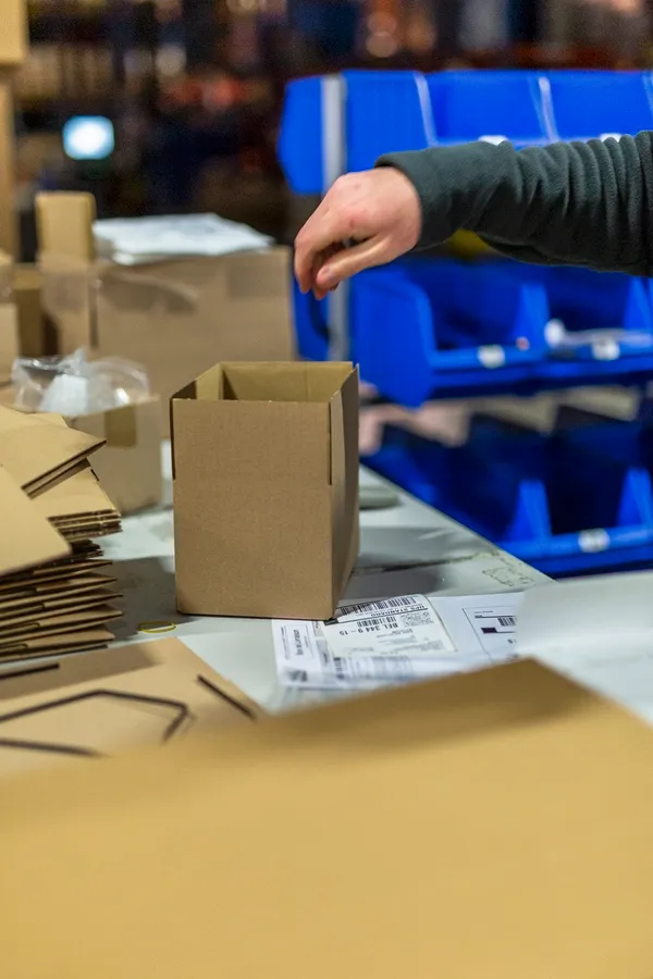 Carton ouvert sur une table de préparation, casiers bleus et étiquette d’expédition.